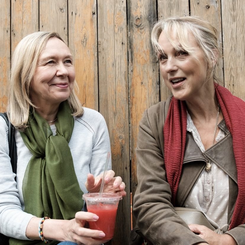 Two mature women are sitting on a bench drinking a juice. They are chatting and having a break.