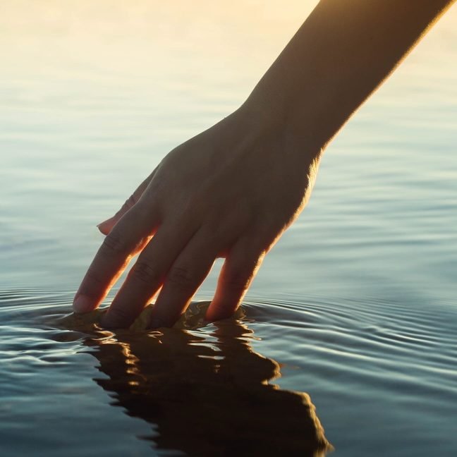 A female hand touching the ocean water in front of a beautful sunset during summer time.
